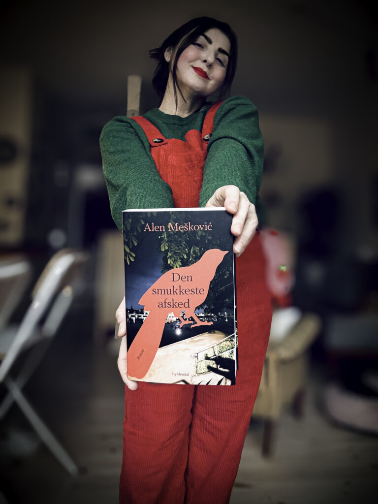 Kvindelig anmelder iført grøn bluse og røde smækbukser holder bogen “Den smukkeste afsked” af Alen Mešković frem mod kameraet.